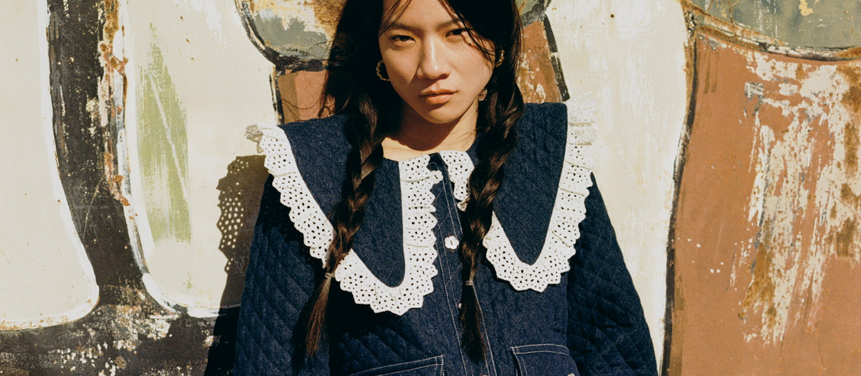A woman wears a denim jacket with lace details leaning against a mural.