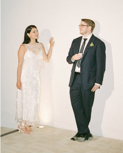 A bride and groom look at one another while standing against a plain wall.