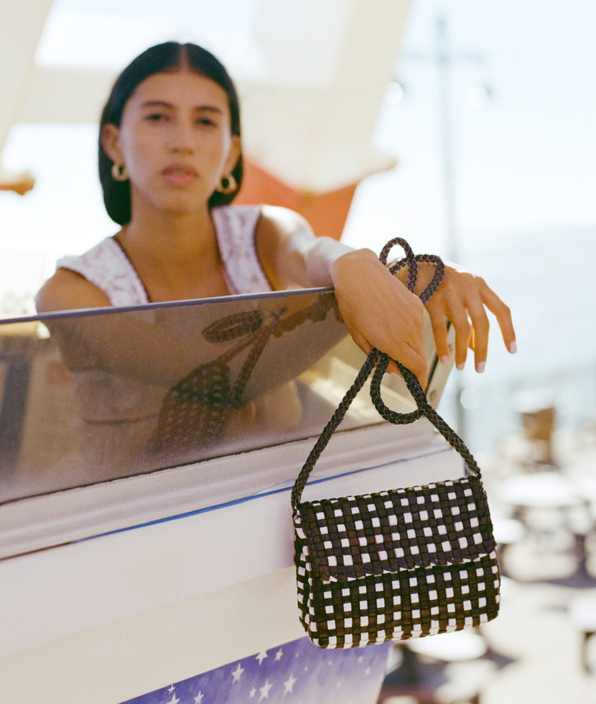 A model leans over a railing holding a brown and white checkered crossbody.