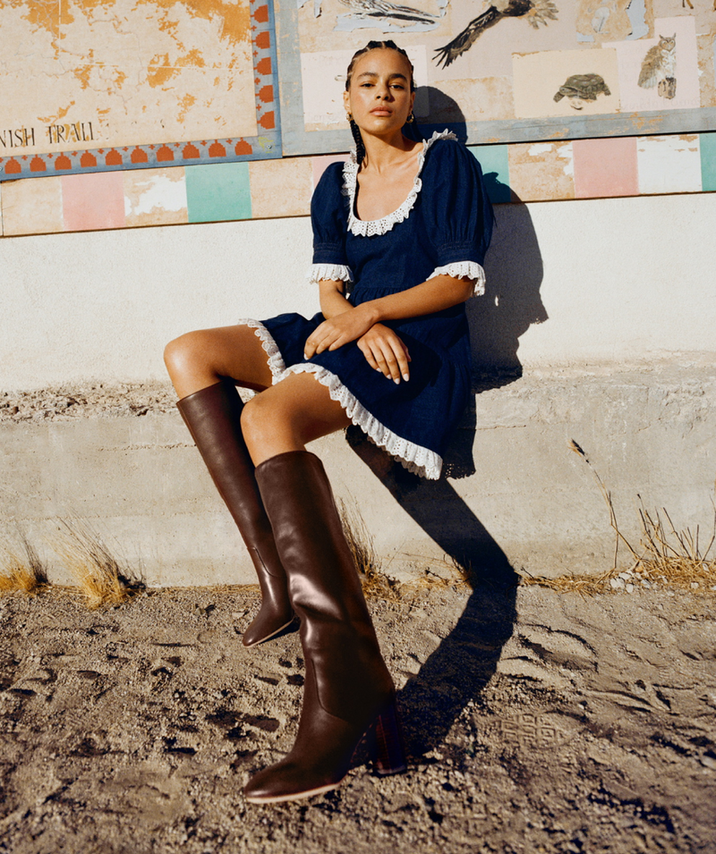 A woman in a blue dress with white lace accents and brown knee-high leather boots sitting against a wall with a mural.