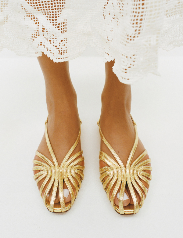 Gold lattice style open-toe flat sandals worn with a white lace dress on a white background.