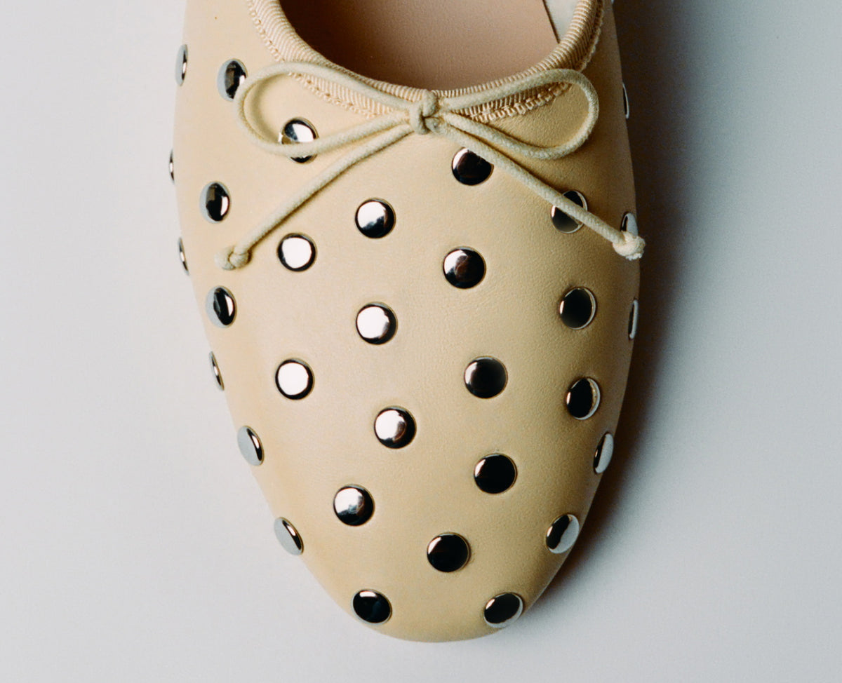 A close up of the toe of a ballet flat in soft yellow with silver studs on a grey background.