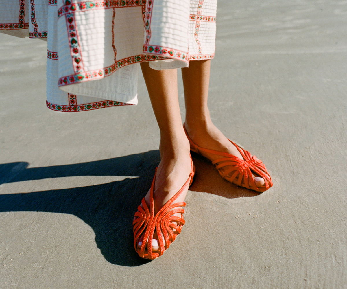 A model stands in the sand wearing a pair of red lattice sandals and a red and white skirt.
