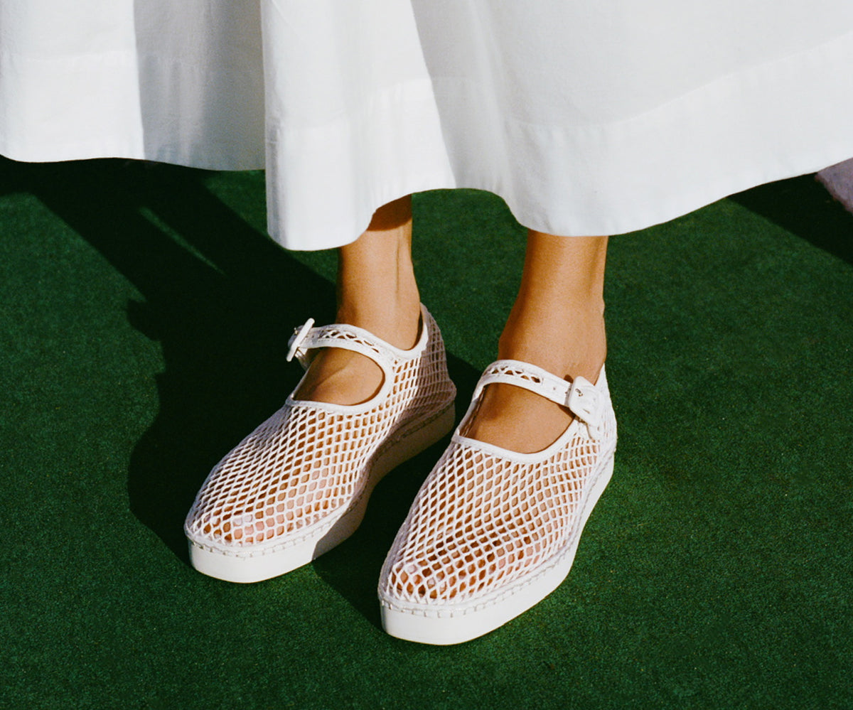 A close up of a model's feet standing on green grass wearing white mesh mary jane sneakers and a white dress.