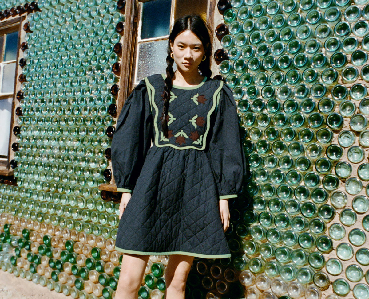Woman in a dark dress with green tulips on the chest standing in front of a wall made of green glass bottles.