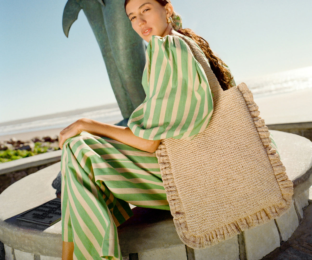 A model wears a green striped set with a natural crochet tote sitting on the ledge of a fountain.
