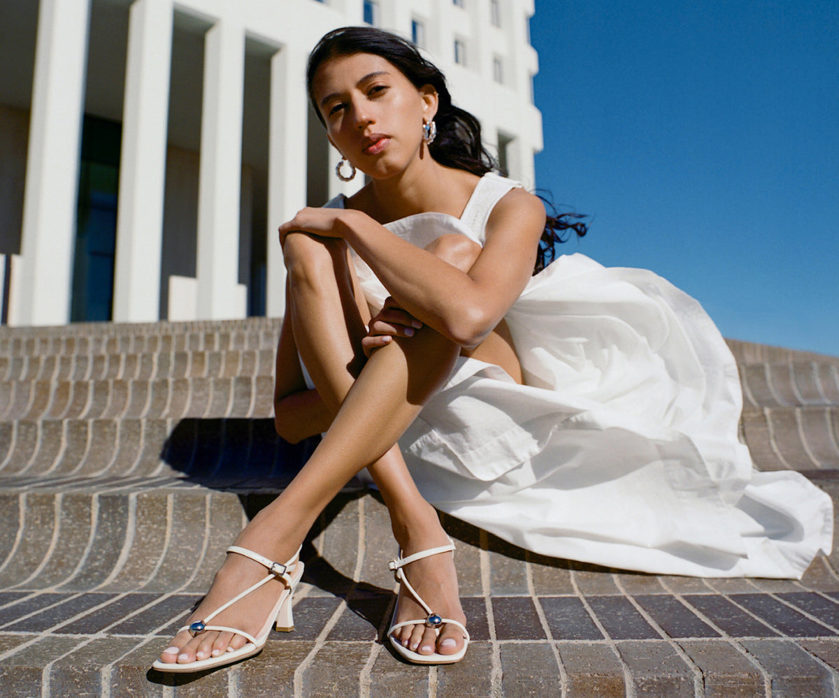 A model sits on a brick sidewalk wearing a white dress and a pair of white strappy heeled sandals.