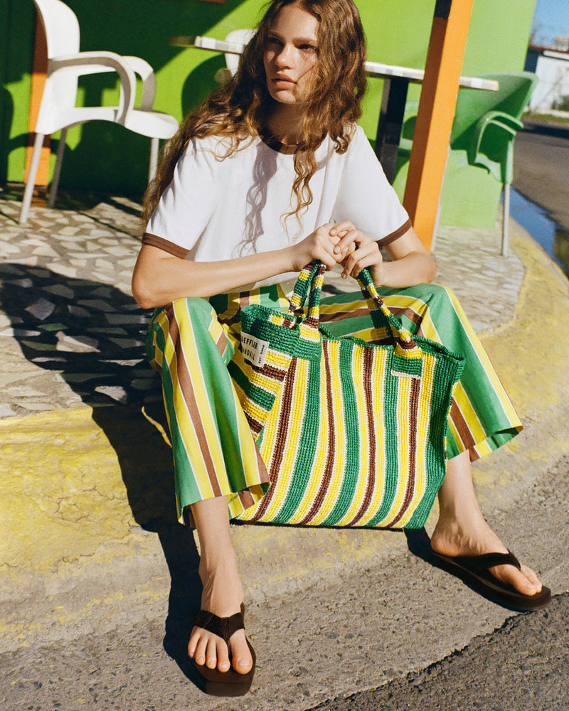 Harly Tee in white cotton, Pasha Trouser in green/yellow/brown striped cotton-linen, Chase Brown Flip Flop, and Sol Green/Yellow Crochet Beach Tote.